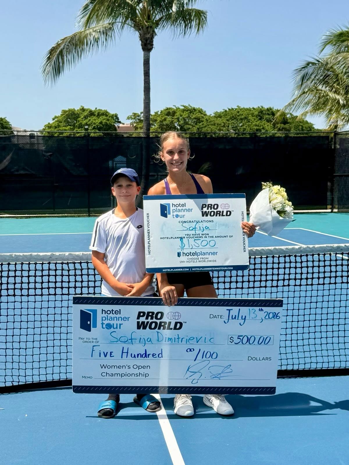 Two tennis players on a court pose with a large ceremonial check and flowers after winning a championship.
